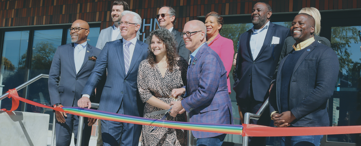 Photo of a group of business men and women smiling and cutting a rainbow ribbon.