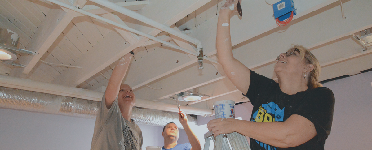 A photo of three people, smiling and reaching up to the rafters on the ceiling with paint brushes.