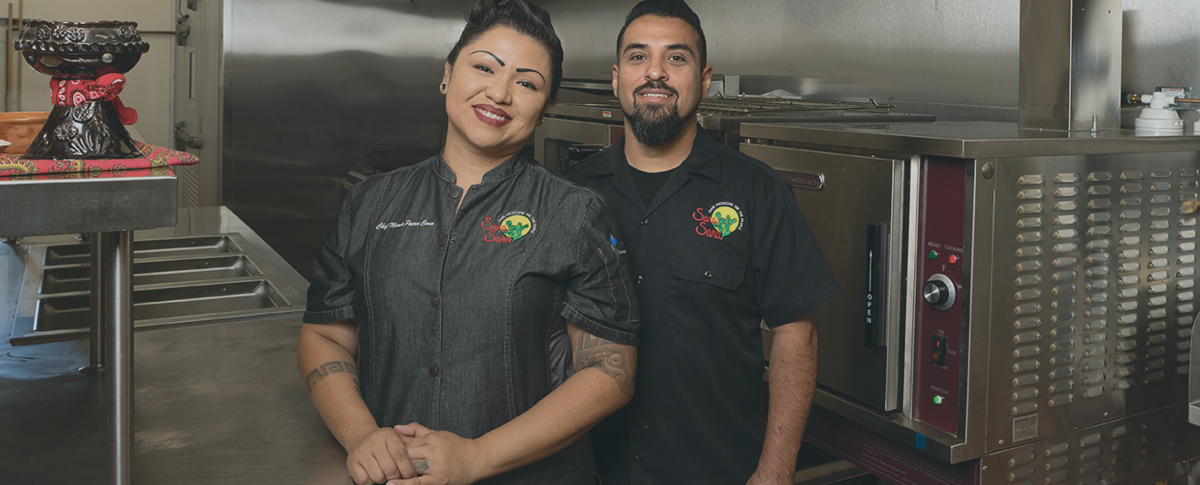 Two people smiling and facing the camera. They are wearing matching black restaurant uniforms and appear to be in a commercial kitchen. 