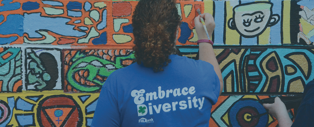 Woman with brown hair facing away from the camera, painting a mural wearing a blue shirt that says "embrace diversity"